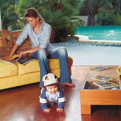 Criança com capacete a engatinhar em sala com mulher no sofá amarelo e mesa de centro com livros e piscina ao fundo.