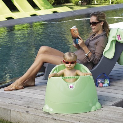 Mulher sentada numa espreguiçadeira junto à piscina lendo e bebé em assento verde claro para bebé.