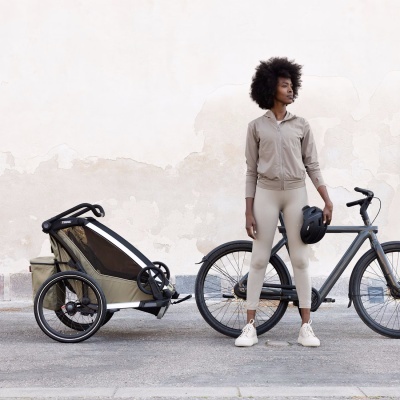 Mulher com roupa desportiva ao lado de bicicleta preta e atrelado Thule verde-oliva