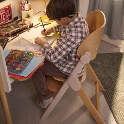 Cadeira infantil de madeira com assento curvo num quarto de estudo com materiais de pintura.