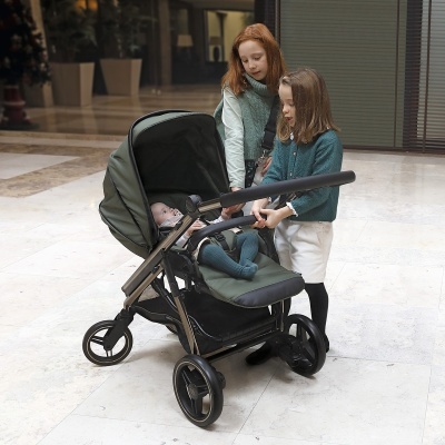 Carrinho de bebé verde com duas crianças em ambiente interior.