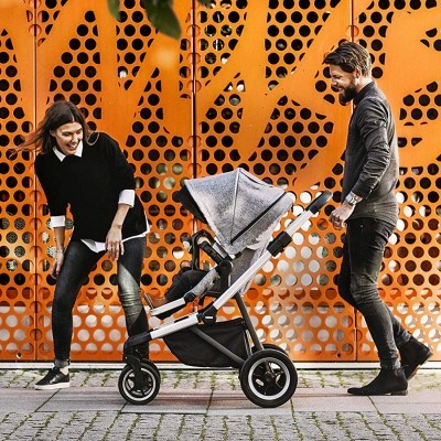 Carrinho de bebé cinza com rodas pretas e jantes brancas ao lado de painel laranja perfurado.