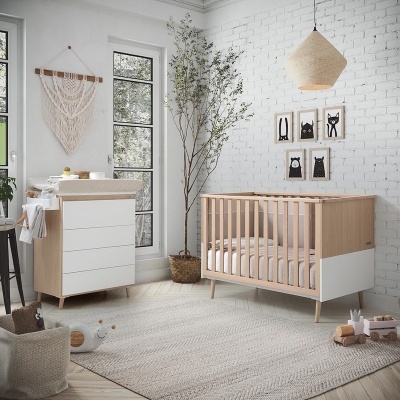Quarto infantil com berço e cómoda em madeira clara e branca, tapete bege, árvore em vaso e decoração de parede