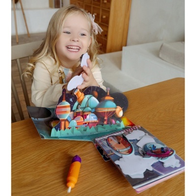 Criança a brincar com livro pop-up de balões de ar quente e caneta laranja numa mesa de madeira