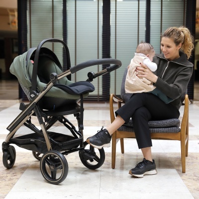 Carrinho de bebé verde escuro com estrutura preta ao lado de mulher sentada segurando bebé