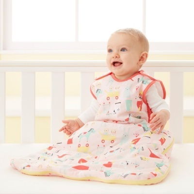 bebé a usar saco de dormir colorido com padrão geométrico e carrinhos sentado numa cama branca