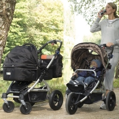 Dois carrinhos de bebé, um com capota fechada preta e outro castanha com criança, ao ar livre com árvores e mulher a pé.