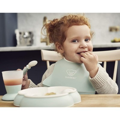 Criança com babete verde-água sentada à mesa a segurar colher e copo