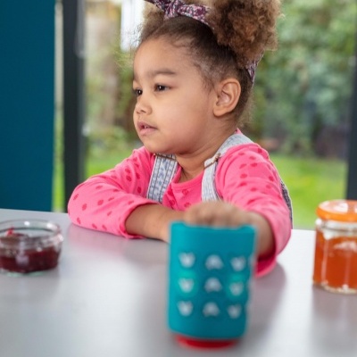 Criança com camiseta rosa e jardineira azul sentada a mesa branca com copo azul e frascos de compota
