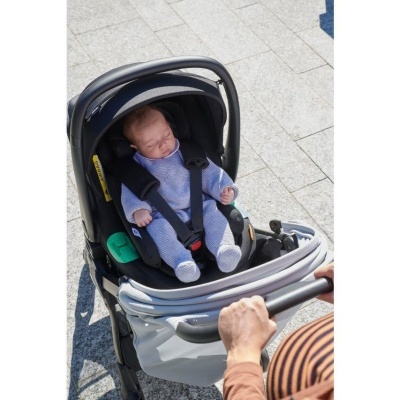 Bebé a dormir na cadeira auto preta com arnês e fato azul, num carrinho cinzento ao ar livre
