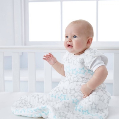 Bebé num saco de dormir branco com estrelas cinzentas e azuis sentado numa cama branca perto de uma janela