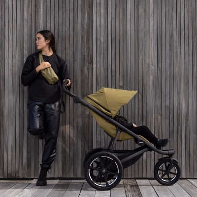 Carrinho de bebé preto com tecido verde-oliva e mulher com roupa preta encostada a parede de madeira