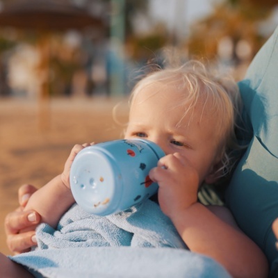 Criança a beber de um copo azul com manchas vermelhas, envolta em toalha azul, segurada por adulto com camisa azul