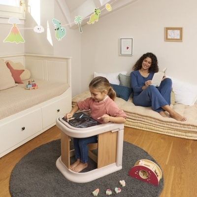 Criança a desenhar numa secretária branca e madeira clara com tampo de ardósia num quarto com cama e mulher sentada em colchão