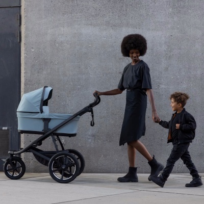 Carrinho de bebé azul claro com estrutura preta empurrado por mulher com vestido preto e criança ao lado numa rua com parede cinzenta.