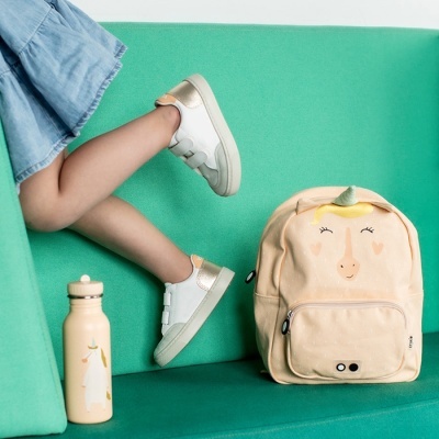 Sapatilhas brancas e douradas, mochila bege com unicórnio e garrafa bege com unicórnio num sofá verde