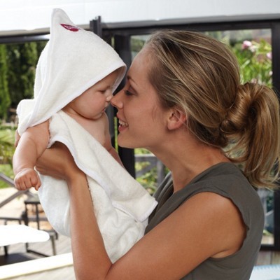 Bebé com toalha branca com capuz segurado por mulher dentro de casa com janelas grandes