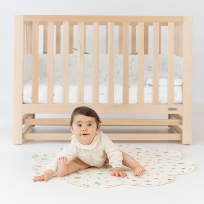Berço de madeira clara com bebê sentado em tapete com padrões coloridos
