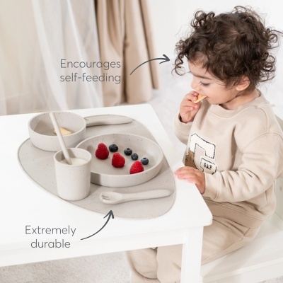 Conjunto de alimentação infantil cinzento com prato, copo, colher e esteira sobre mesa branca, criança a comer