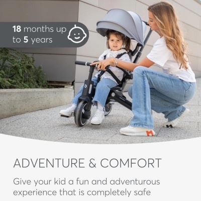 Criança num triciclo preto com cobertura cinzenta, ao ar livre com acompanhante.