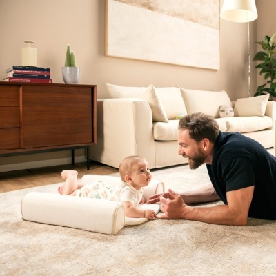 Bebé deitado em almofada creme com apoio acompanhado por homem de camisa preta numa sala com tapete bege, sofá creme, cómoda de madeira e candeeiro branco.