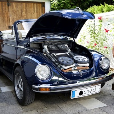 Carro conversível clássico azul com capô aberto mostrando motor elétrico e malas ao lado