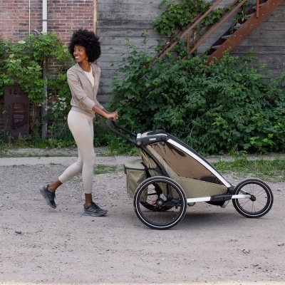 Mulher empurra carrinho de bebé desportivo bege e branco ao ar livre em ambiente urbano com vegetação