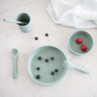 Utensílios infantis de silicone verde claro com frutas sobre mesa clara