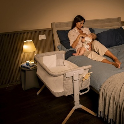 Berço portátil cinza junto a cama com mulher amamentando bebé e luminária amarela em mesa ao lado