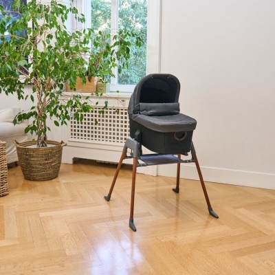 Berço portátil cinzento com pernas em madeira clara, junto a planta em vaso de verga