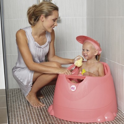 Banheira cor-de-rosa com bebê usando boné cor-de-rosa em casa de banho e mulher a interagir.