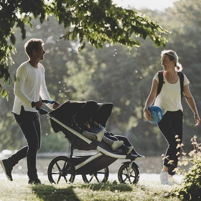 Carrinho de bebé duplo preto e cinza a ser empurrado em parque ensolarado