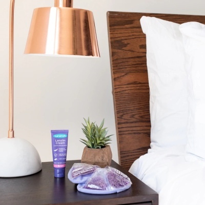 Crado-mudo com lâmpada de metal cobre, tubo roxo e pantufas lilás ao lado da cama com cabeceira de madeira.