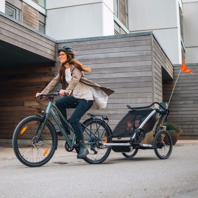 Bicicleta verde-acinzentada com reboque para criança bege e capacete preto numa rua urbana