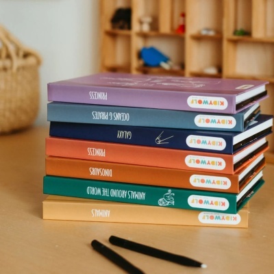 Seis livros infantis variados empilhados sobre uma mesa de madeira clara com dois marcadores pretos ao lado.