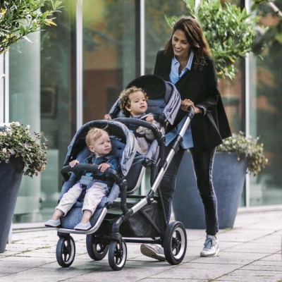 Carrinho de bebé duplo cinza com mulher a empurrá-lo numa calçada ao ar livre