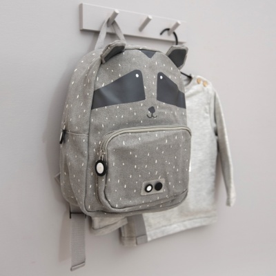 Mochila infantil cinzenta com rosto de guaxinim pendurada numa parede cinza ao lado de uma camisa cinza clara