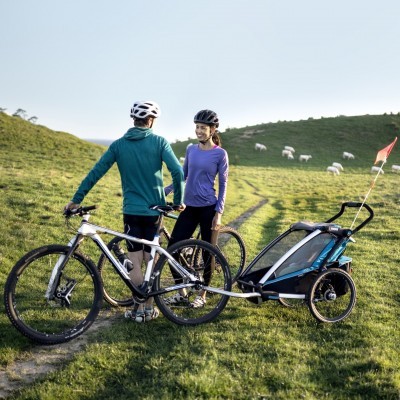 Casal com bicicletas e atrelado azul num campo verde com ovelhas de fundo
