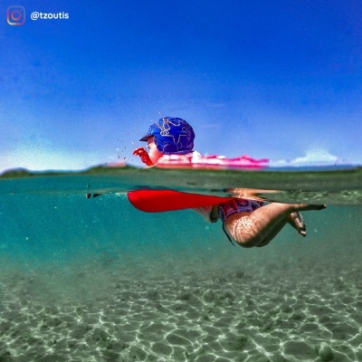 Criança a nadar com colete salva-vidas vermelho e boné azul em água cristalina sob céu azul