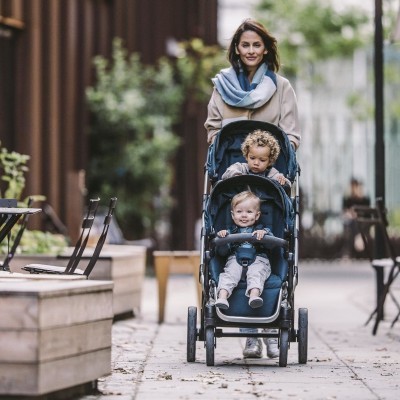 Carrinho duplo de bebé azul-escuro com crianças e mulher a empurrar na rua ao ar livre