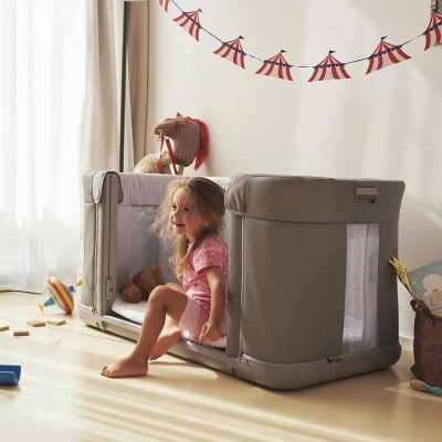 Berço portátil cinza com criança sentada à borda e decoração de bandeirolas vermelhas e azuis na parede