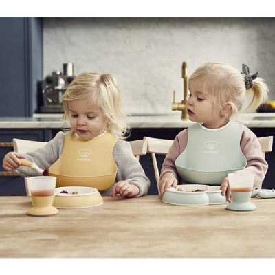 Duas crianças pequenas com babetes de silicone e pratos e copos da mesma cor numa mesa de cozinha.