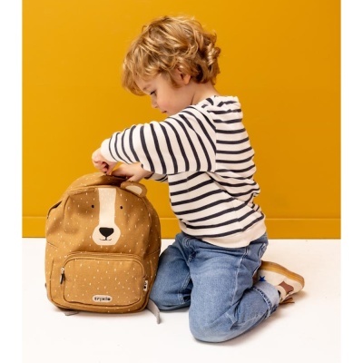 Mochila castanha com rosto de urso ao lado de criança com camisa às riscas e calças jeans contra fundo amarelo