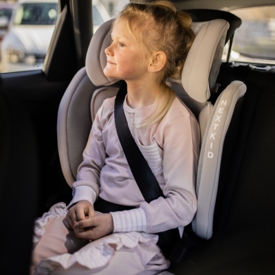 Cadeira auto cinzenta clara com espuma branca e texto NEXT KID, menina loira sentada no banco traseiro do carro