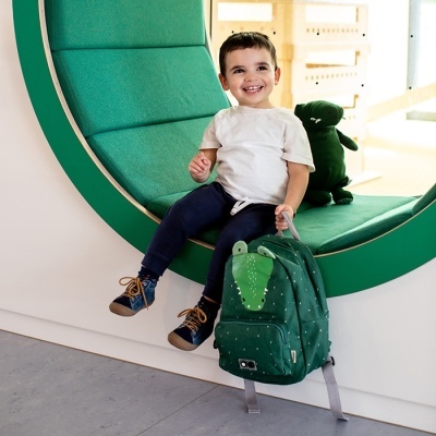 Criança com mochila verde de dinossauro sentada em banco verde oval