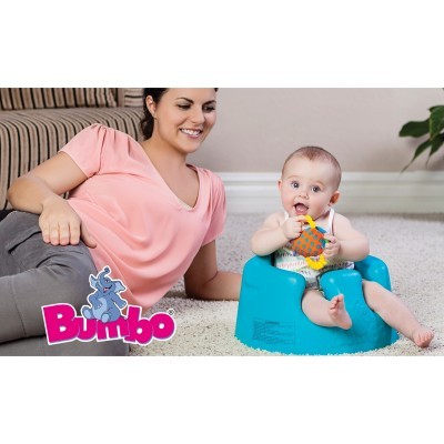 Bebé sentado em cadeira azul Bumbo com brinquedo, mulher sorridente ao lado numa sala clara com tapete