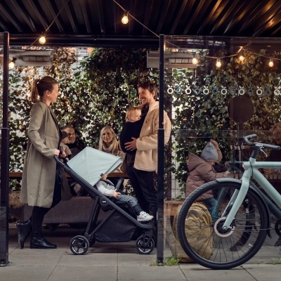 Carrinho de bebé cinza claro com estrutura preta, criança sentada, adultos num café exterior coberto com bicicleta à direita