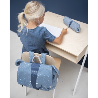 Mochila azul clara com padrão de pontos brancos numa cadeira, criança a escrever numa secretária