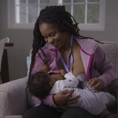 Mulher a amamentar bebé num sofá cinzento com casaco rosa e fita laranja ao pescoço