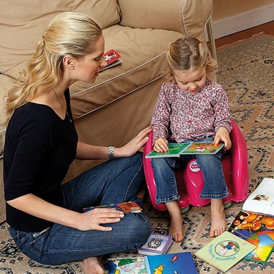 Menina numa cadeira infantil rosa a ler um livro, com livros espalhados no tapete e uma mulher ao lado.